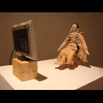 traditional plains First Nations doll seated in a chair in front of a TV monitor